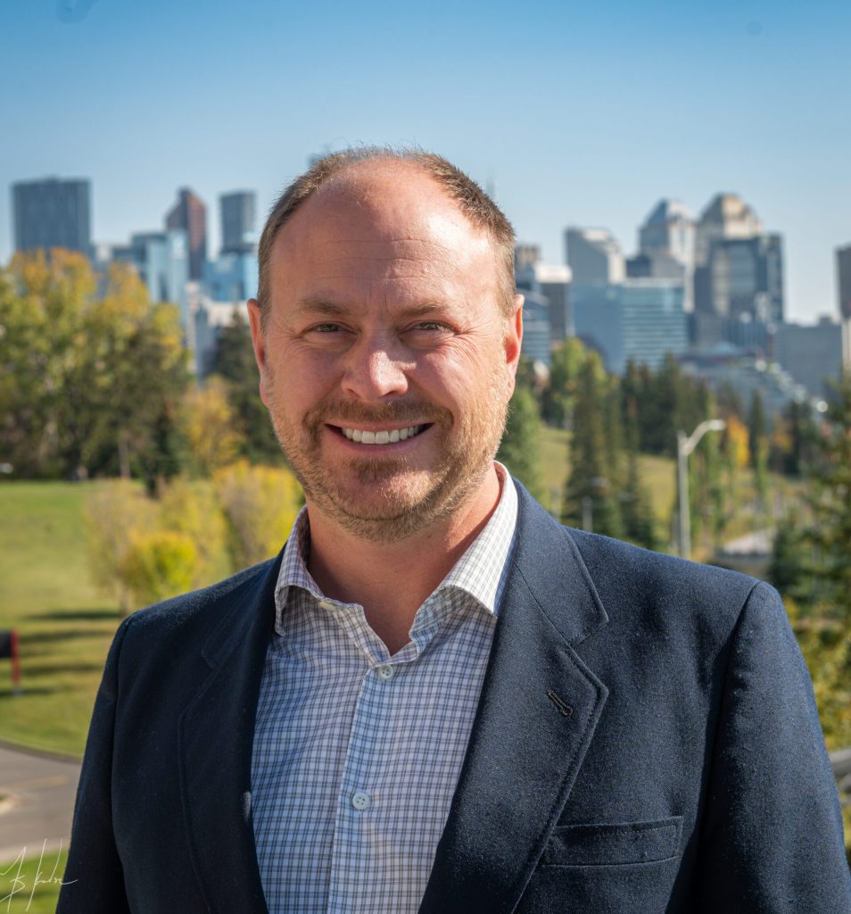 A photo of Tyler Nagel in front of a cityscape wearing a dark blue blazer.